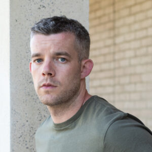 Portrait of the actor Russell Tovey. Russell is a white male with short grey hair, a dusting of stubble on his chin and is wearing a green grey top. He is stood in front of a cream coloured brick wall. 